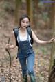 A woman in overalls and a backpack holding a stick in the woods.